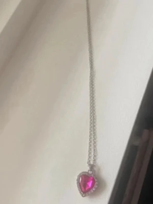 Halsband med hjärthänge i rosa sten - Snyggt halsband med ett hjärtformat hänge i klarrosa sten, omgiven av små gnistrande stenar. Kedjan är silverfärgad och smal, vilket ger ett stilrent intryck. Perfekt accessoar för att lyfta din look med lite extra färg och bling.
