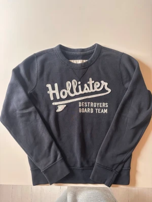 Mörkblå Hollister sweatshirt - Mörkblå sweatshirt från Hollister med broderad text 'Hollister Destroyers Board Team' på bröstet. Tröjan har rund halsringning, långa ärmar och ribbade muddar. Perfekt för en avslappnad och sportig stil.