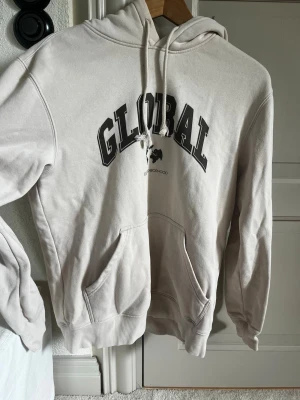 Beige hoodie med tryck GLOBAL - Snygg beige hoodie med huva och dragsnören. På bröstet står det 'GLOBAL' i stora svarta bokstäver och ett mindre tryck under. Hoodien har känguruficka och ribbade muddar vid ärmar och nederkant. Perfekt för en chill och avslappnad stil.
