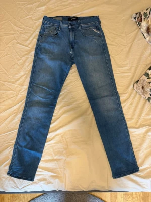  Replay Anbass jeans - Snygga blå slim fit jeans från Replay med klassisk femficksdesign och ljus tvätt. Jeansen har normal midja, dragkedjegylf och detaljerade sömmar. Perfekta för en avslappnad och trendig stil. På bild 2 ser man ett litet slitage, det är inget hål utan bara ett litet slitage. Så absolut inget stort så. Som man även ser på bilderna har förkortat jeansen lite när jag köpte de. Längd 99cm, midjan 35cm. Storlek W29 L32. Vid funderingar eller liknande är det bara att höra av sig!