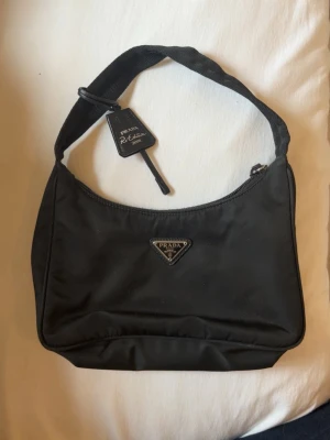 Svart handväska från Prada - (Såklart äkta)  svart handväska från Prada Re-Edition med klassisk trekantig logga framtill. Väskan har en enkel axelrem och är tillverkad i nylonmaterial. Stängs med dragkedja och har en stilren, minimalistisk design.