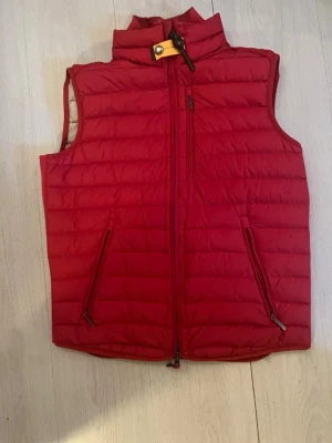 Röd parajumper väst med dragkedjor - Snygg röd dunväst med hög krage och dragkedja framtill. Västen har två snedställda fickor med dragkedja och en bröstficka. Quiltad design och justerbar nederkant. Perfekt för lager-på-lager-stil.