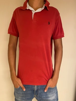 Röd pikétröja från Polo Ralph Lauren - Klassisk röd pikétröja från Polo Ralph Lauren med vit krage och knappslå. Tröjan har korta ärmar och den ikoniska loggan broderad på bröstet. Perfekt för en avslappnad och stilren look.