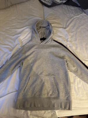Grå hoodie från GANT med detalj på sidorna - Snygg grå hoodie från GANT med huva och känguruficka framtill. Tröjan har en röd och marinblå rand längs ena ärmen som ger en sportig vibe. Tillverkad i mjukt bomullsmaterial och har diskret GANT-logga på fickan. 