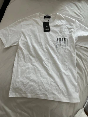 Amiri T-shirt - Säljer en vit t-shirt från AMIRI med coolt tryck framtill och stort AMIRI-tryck på ryggen med färgstänk och droppande effekt. T-shirten har rund hals och korta ärmar. Perfekt för dig som gillar streetwear och vill sticka ut.