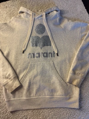 Isabelle marant hoodie - Snygg isabel marant hoodie med stor ficka framtill och huva med dragsko. På bröstet finns ett stort ljusblått glittertryck med texten 'marant' och en grafisk logga. Hoodien har ribbade muddar vid ärmar och nederkant. Perfekt för en chill och trendig look.
