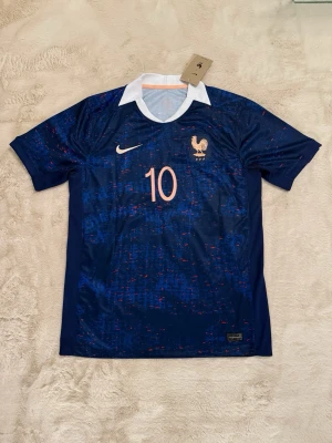 Frankrike fotbollströja Mbappé #10 - Mörkblå fotbollströja från Nike med Frankrikes landslagsemblem och nummer 10 på bröstet. Baksidan har namnet Mbappé och stort nummer 10. Tröjan har vit krage och detaljer i ljusrosa samt korta ärmar. Perfekt för dig som älskar fotboll och vill stötta Frankrike.