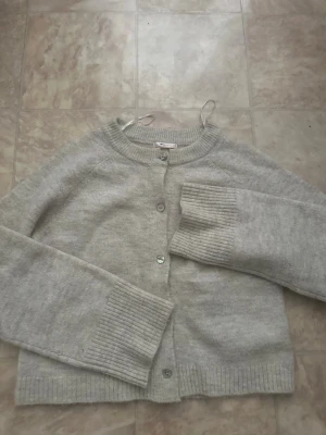 Beige kofta från Gina Tricot - Mysig beige kofta från Gina Tricot med rund halsringning och knäppning framtill. Koftan har långa ärmar och breda ribbade muddar vid ärmslut och nederkant. Perfekt att slänga på sig när det är lite kyligt ute. Helt sprillans ny kofta 🎀. 