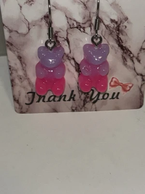 Lila och rosa nalleörhängen - Söta örhängen formade som gummibjörnar i lila och rosa. Hängande modell med metallkrok. Perfekta för dig som gillar lekfulla accessoarer och vill sticka ut med färgglada detaljer. Örhängens krokarna är i rostfritt stål 