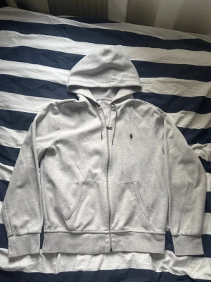 Grå hoodie från Polo Ralph Lauren - Snygg ljusgrå hoodie från Polo Ralph Lauren med dragkedja, två fickor framtill och klassisk huva med dragsko. Ikoniska mörkblå loggan broderad på bröstet. Tillverkad i mjukt bomullsmaterial som känns riktigt skönt mot huden.