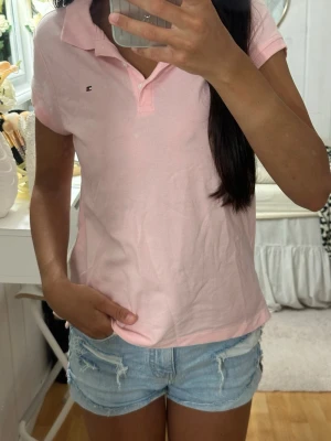 Ljusrosa pikétröja Tommy Hilfiger - Säljer en ljusrosa pikétröja från Tommy Hilfiger med klassisk krage och korta ärmar. Tröjan har en liten logga på bröstet och är tillverkad i mjuk bomull. Perfekt för en avslappnad och stilren look.