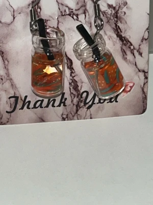 Örhängen med juiceglas och sugrör - Unika örhängen formade som små juiceglas med sugrör. Glasen är genomskinliga med orangefärgad vätska och dekorationer som liknar fruktbitar och blad. Perfekt för dig som gillar lekfulla och färgglada accessoarer. Örhängens krokarna är i rostfritt stål 