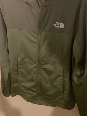Grön vindjacka från The North Face - Snygg vindjacka från The North Face i två nyanser av grönt. Jackan har hel dragkedja framtill, huva och logga på både bröstet och ryggen. Perfekt för blåsiga dagar och enkel att matcha med olika outfits.
