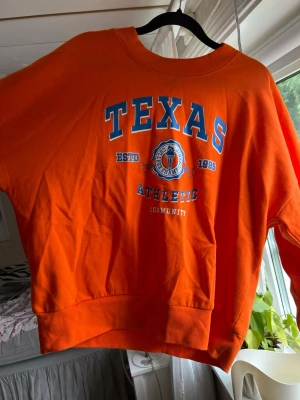 Orange sweatshirt med Texas-tryck - Cool orange sweatshirt med stort blått och vitt Texas-tryck på bröstet. Tröjan har rund halsringning, ribbade muddar och avslappnad passform. Perfekt för dig som gillar college-stil och sportiga vibes.