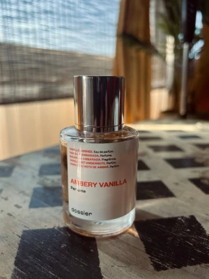 Ambery Vanilla parfym från Dossier 50ml - Ambery Vanilla parfym från Dossier i en rund glasflaska med silverfärgat lock. Doften har inslag av vanilj och amber, och flaskan har en minimalistisk design med vit etikett och röd text. Perfekt för dig som gillar varma och söta doftnoter. Ingen låda.