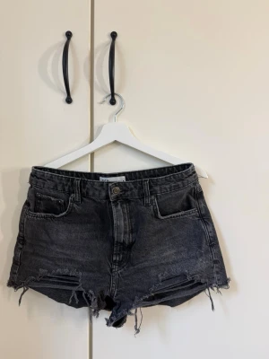 Svarta jeansshorts med fransig kant - Säljer ett par svarta jeansshorts med hög midja och rå, fransig kant. Shortsen har fem fickor och slitna detaljer framtill för en avslappnad look. Från Zara i strl 40