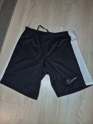 Svarta Nike shorts med vita sidor - Säljer ett par svarta Nike shorts med vita sidopaneler och snörning i midjan. Shortsen har en sportig look med Nike-logga på ena benet och är tillverkade i ett lätt och bekvämt material som passar perfekt för träning.
