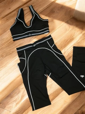 Svart träningsset från Alo - Snyggt svart träningsset från Alo med sport-bh och tights. Setet har vita kontrastsömmar och logga framtill. Materialet är stretchigt och passar perfekt för gym, yoga eller löpning. Sport-bhn har breda axelband och tightsen har hög midja.