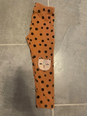 Prickiga leggings med kattmotiv - Snygga bruna leggings med svarta prickar och gulliga kattansikten på knäna. Mjukt och stretchigt material som sitter skönt. Perfekta för dig som gillar lekfulla detaljer och vill sticka ut lite extra.