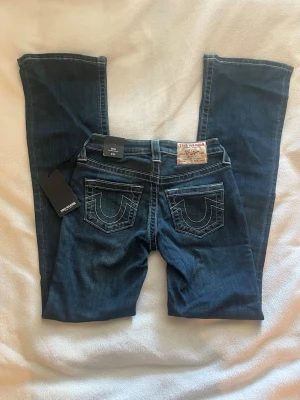 Mörkblå True Religion jeans bootcut - Snygga mörkblå jeans från True Religion med bootcut-modell och klassiska kontrastsömmar. Bakfickorna har märkets ikoniska broderi och läderpatch i midjan. Perfekta för dig som gillar en tidlös och cool stil. Modellen heter BECCA mid rise bootcut 🔥storlek: W23, Innerbenslängd 83,5cm 🌟nyköpta och aldrig använda, kommer med prislapp ⚡️⚡️