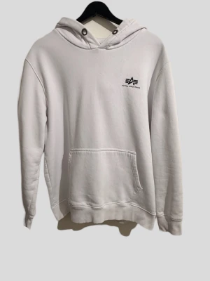Vit hoodie från Alpha Industries - Snygg vit hoodie från Alpha Industries med liten logga på bröstet. Klassisk känguruficka framtill och huva med dragsko. Tillverkad i mjukt material som känns skönt mot huden. Perfekt för en avslappnad och stilren look.