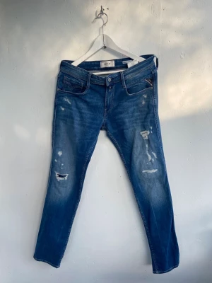 Blå Repley jeans - Säljer ett par blå Replay jeans med slitningar och distressed detaljer på framsidan. Jeansen har fem fickor, normal midja och klassisk dragkedja med knapp. Materialet är denim i bomull med lite stretch för skön passform. Använda fåtal gånger. Storlek 30/32