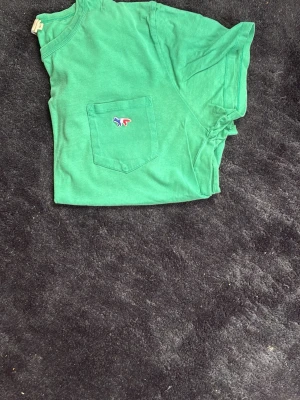 Grön t-shirt med ficka från Maison kitsune - Snygg grön t-shirt med rund hals och en bröstficka som har en liten broderad räv i flera färger. T-shirten har korta ärmar och är gjord i mjukt bomullstyg. Perfekt för en avslappnad stil och enkel att matcha med jeans eller shorts.