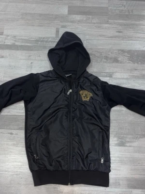 Svart hoodie med dragkedja och emblem - Snygg svart hoodie med dragkedja och huva. Framsidan har ett guldfärgat emblem på bröstet och två fickor med dragkedja. Ärmar och baksida är i mjukt tyg medan framsidan har en glansigare finish. Perfekt för en chill och stilren look.