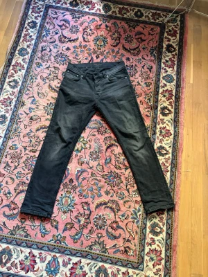 Svarta Nudie Grim Tim jeans  - Säljer ett par svarta slim fit jeans med klassisk femficksmodell och normal midja. Jeansen har raka ben och är tillverkade i ett stretchigt material för extra komfort. Passar perfekt till en avslappnad och trendig stil. W31 L30