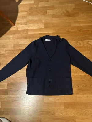 Mörkblå kavaj med fickor - Stilren mörkblå kavaj med tre knappar framtill och två stora framfickor. Klassisk krage och rak passform. Perfekt för en snygg och enkel look. Materialet ser ut att vara mjukt och bekvämt, troligen bomull eller en bomullsblandning.