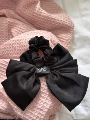 Svart scrunchie med stor rosett - Snygg svart scrunchie från ghd med en stor, elegant rosett. Perfekt för att ge frisyren en extra touch och hålla håret på plats. Passar både till halvuppsättning och helt uppsatt hår.