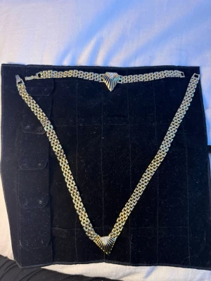 Halsband och armband set - Vintage smyckesset från 80-talet. Snyggt halsband och armband i guldfärgad metall med bred, platt länk och ett dekorativt geometriskt mönster i mitten. Smyckena är osignerade (saknar stämplar), vilket var vanligt för många mode- och bijouterismycken under denna era. Tung och gedigen kvalitet som känns lyxig att bära. Perfekt till festen, nyår eller för att liva upp vardagslooken!