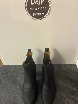 Svarta Chelsea boots i skinn - Säljer ett par klassiska svarta Chelsea boots i skinn med rundad tå och resår på sidorna. Insidan är fodrad i brunt material och bootsen har dragflikar baktill för enkel påtagning. Perfekta för en stilren och tidlös look.