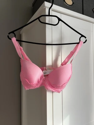 Rosa push-up bh med spetsdetaljer - Söt rosa push-up bh med vadderade kupor och fina spetsdetaljer längs nederkanten. Justerbara axelband och hakknäppning bak för bästa passform. Perfekt för dig som vill ha både komfort och en feminin look.
