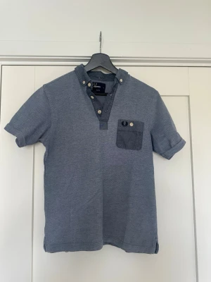 Blå henley-piké med knappdetaljer - Snygg blå henley-pike med korta ärmar och kontrasterande bröstficka med knappdetaljer. Skjortan har en klassisk krage med knappar och en avslappnad passform. Perfekt för en casual look och enkel att matcha med jeans eller shorts. Storlek s 