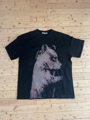 Acne studios t shirt - Säljer en svart t-shirt från Acne Studios med ett stort tryck av en björn på framsidan. T-shirten har rund hals och korta ärmar. Materialet ser ut att vara mjuk bomull, perfekt för en avslappnad och cool stil.