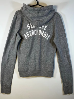 Abercrombie & Fitch Grå Zip Hoodie M – New York Logo - Grå Abercrombie & Fitch zip hoodie i storlek M. Klassisk äldre A&F-modell med broderad vit älg på bröstet och stort New York Abercrombie-tryck på ryggen.  Snygg melerad grå färg, full zip, huva och två fickor framtill. Perfekt för en klassisk vintage/Y2K Abercrombie-look.  Begagnat men fint skick, nån liten ljus missfärgning bredvid dragkedjan men inget jättetydligt, se bilder.