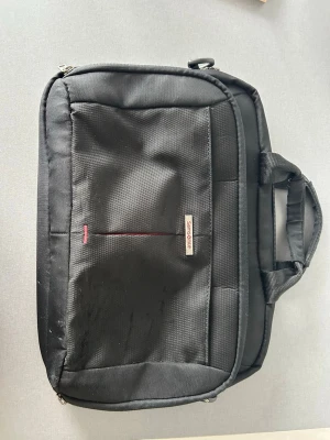 Svart datorväska med handtag - Samsonite - Snygg och praktisk svart datorväska i slitstarkt tyg med dubbla handtag och dragkedja. Väskan har en enkel design med diskreta röda detaljer och en liten logotyp på framsidan. Perfekt för att bära din laptop till skolan eller jobbet.