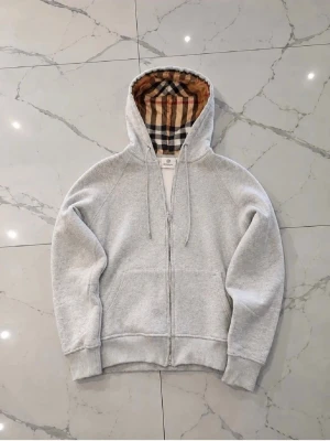 Grå hoodie med rutigt foder i huvan - Snygg ljusgrå Burberryhoodie med dragkedja och känguruficka. Huvan är fodrad med ett klassiskt beige, svart och röd-rutigt tyg som ger en unik touch. Tröjan har ribbade muddar vid ärmar och midja samt dragsko i huvan. Perfekt för en chill och stilren look.