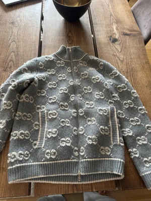 Grå stickad ziptröja från Gucci - Snygg grå stickad ziptröja från Gucci med vit GG-logga över hela tröjan. Tröjan har hög krage, dragkedja framtill och ribbade muddar. Två fickor framtill med vita detaljer. Tillverkad i Italien, perfekt för dig som gillar exklusiv streetstyle.