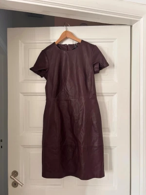 Vinröd skinnklänning med kort ärm - Vinröd klänning i skinn med rund halsringning och korta ärmar. Klänningen har en figurnära passform och dragkedja i ryggen. Stilsäker och enkel design som ger en cool look. Helt ny❤️