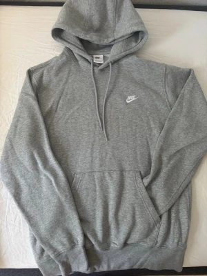 Grå hoodie från Nike med huva - Säljer en klassisk grå hoodie från Nike med vit logga på bröstet. Hoodien har huva med dragsko, stor magficka och ribbade muddar vid ärmar och nederkant. Perfekt för en chill och sportig look.