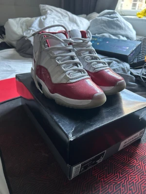 Nike Air Jordan 11 röda och vita - Nike Air Jordan 11 sneakers i vitt och rött med högt skaft. Skorna har en blank röd lackdetalj runt om, vit mesh på ovandelen och en tydlig Jumpman-logga på sidan. Sulan är vit med genomskinliga partier och skorna har vita skosnören.