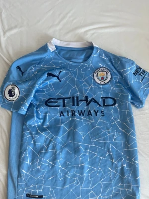 Manchester City matchtröja blå Puma - Säljer en ljusblå Manchester City matchtröja från Puma med vitt grafiskt mönster, klubbmärke på bröstet och Premier League-märke på ärmen. Tröjan har rund hals och sponsorlogga från Etihad Airways på framsidan. Perfekt för fotbollsfans!