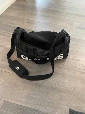 Svart träningsväska från Adidas - Snygg svart sportbag från Adidas med stort vitt logotryck på sidan. Väskan har justerbar axelrem med vadderad axelkudde och två bärhandtag. Perfekt för gymmet eller träningen, tillverkad i slitstarkt material och har dragkedja upptill.