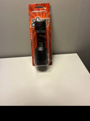 Svart adapter i förpackning - Säljer en svart adapter i hårdplast, fortfarande i originalförpackning med röd bakgrund. Adaptern har en robust form och verkar vara avsedd för verktyg eller maskiner. Perfekt för dig som behöver extra tillbehör till din utrustning.