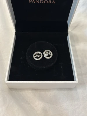 Silverörhängen Pandora med LOVE-text - Säljer ett par runda silverörhängen från Pandora, som inte finns längre att köpa. Materialet är jag osäker, men tror det är gjort av sterlingsilver såsom resten av pandoras silver smycken. Fick dessa i present men insåg väldigt snabbt att det tyvärr var inget för mig, därav är dem helt oanvända. Lådan på bilden medföljer vid köpet☺️