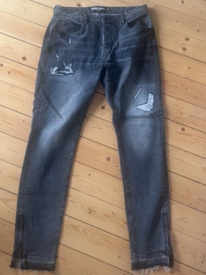 Svarta slitna skinny jeans med dragkedja - Säljer ett par svarta skinny jeans med slitningar och råa kanter nertill. Jeansen har dragkedjor vid bensluten och en tvättad look som ger en cool vibe. Perfekta för dig som gillar en edgy stil. Materialet är klassisk denim.