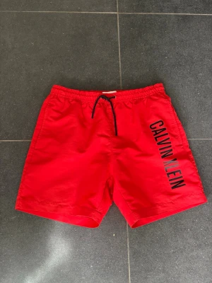 Röda badbyxor från Calvin Klein - Snygga röda badbyxor från Calvin Klein med svart dragsko i midjan och stort logotryck längs ena benet. Klassisk passform med elastisk midja och en bakficka. 