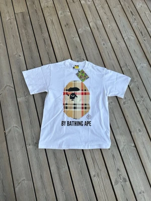 Vit BAPE t-shirt med rutigt tryck - Vit t-shirt från BAPE med stort rutigt apansikte-tryck på bröstet och texten 'BY BATHING APE' under. Klassisk passform och rund hals. Perfekt statement-plagg för streetwear-stilen.